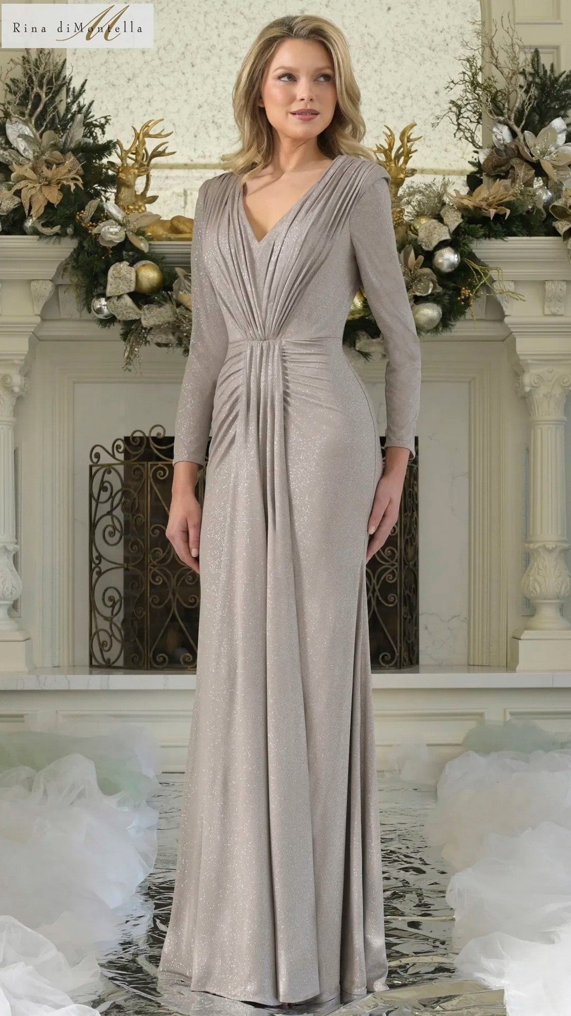 A woman stands indoors in front of a festive mantel wearing the Rina Di Montella RD3348 long formal dress, a taupe, floor-length gown with long sleeves and gathered detailing.